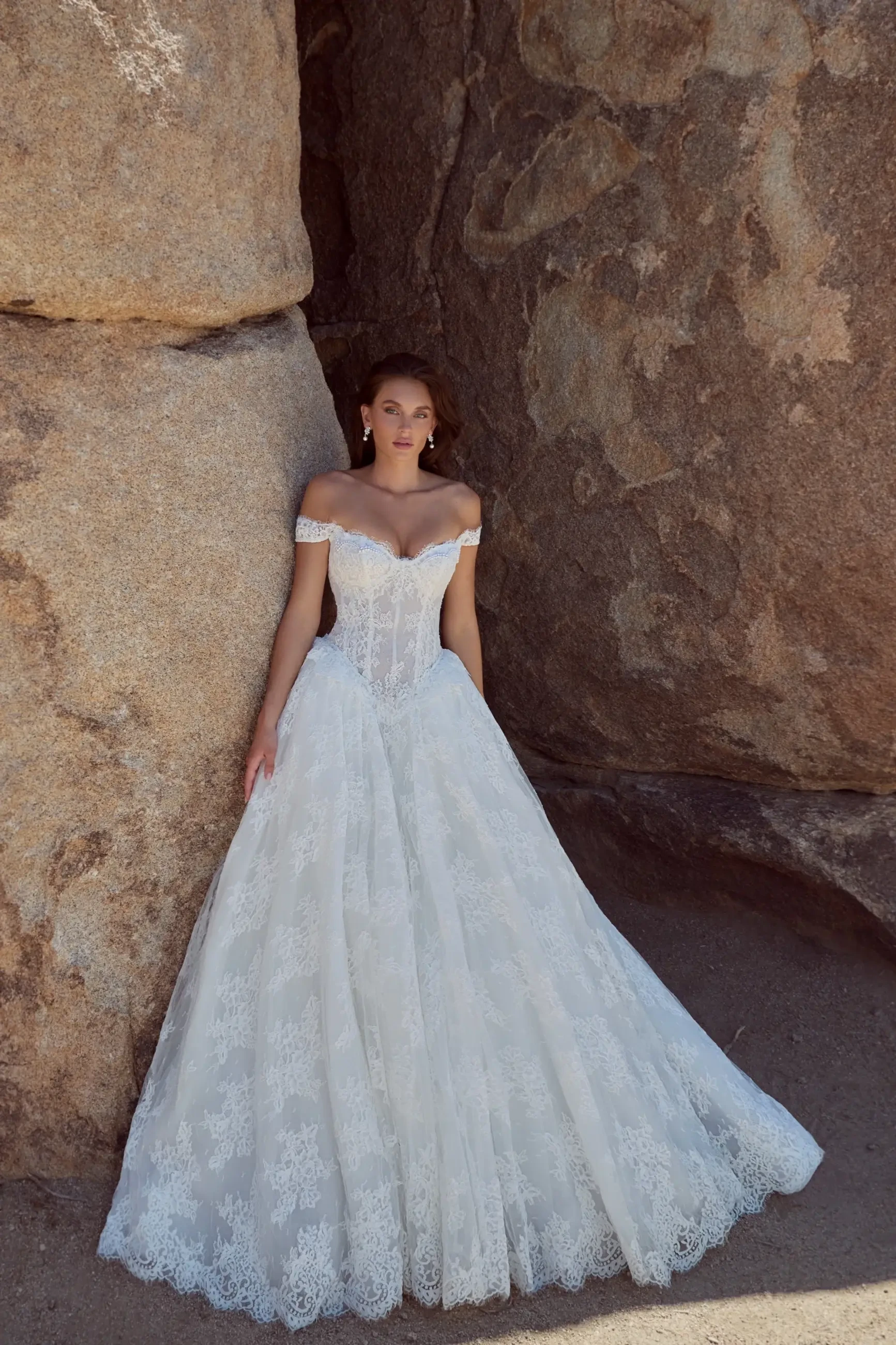A woman in a white, off-the-shoulder wedding dress standing beside large rocks.