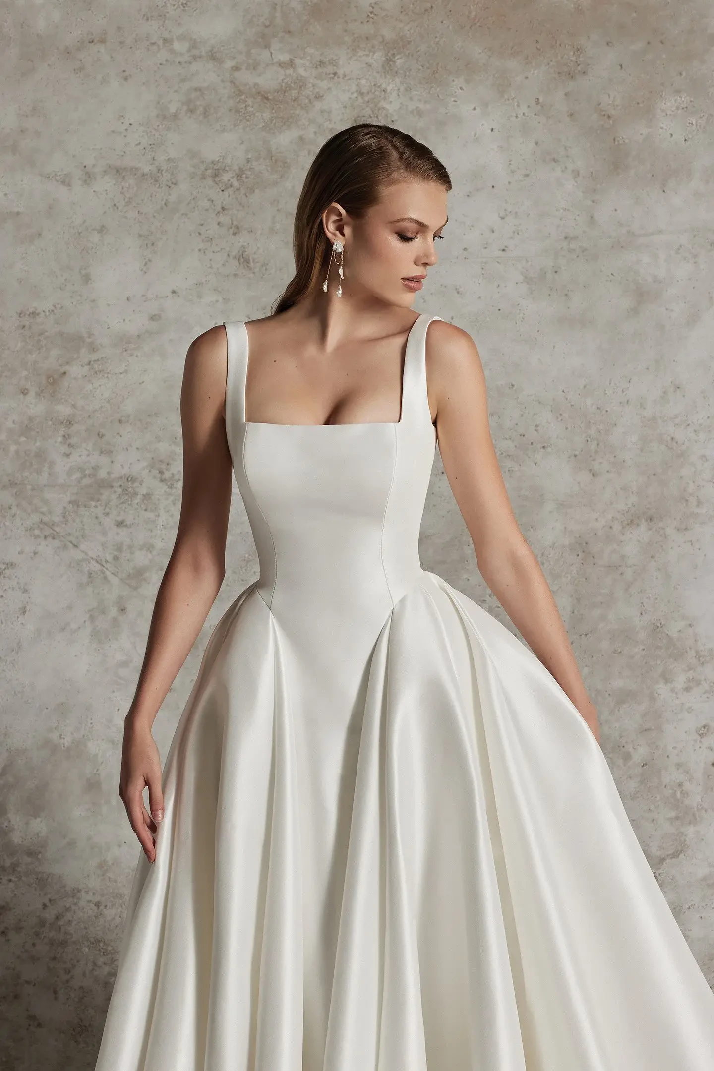 Woman in elegant white gown with a square neckline and full skirt, standing against a textured gray background. She wears drop earrings and looks down, exuding grace.
