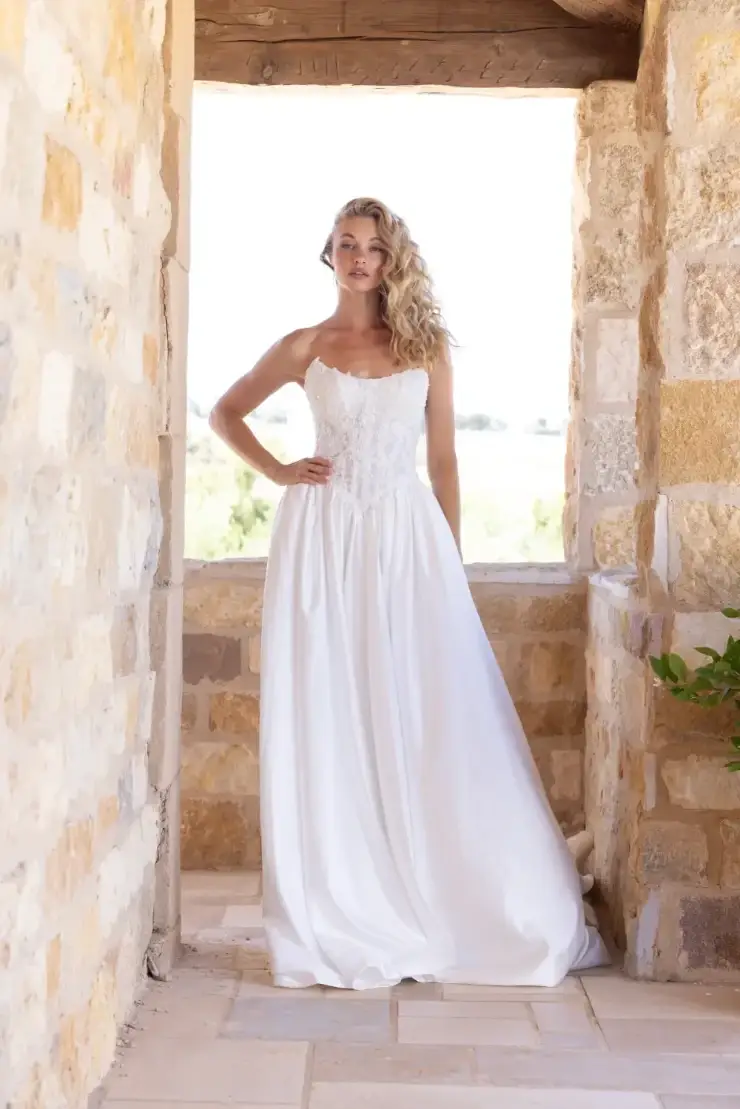 A woman in a white dress standing in an architectural space with stone walls, looking confidently at the camera.