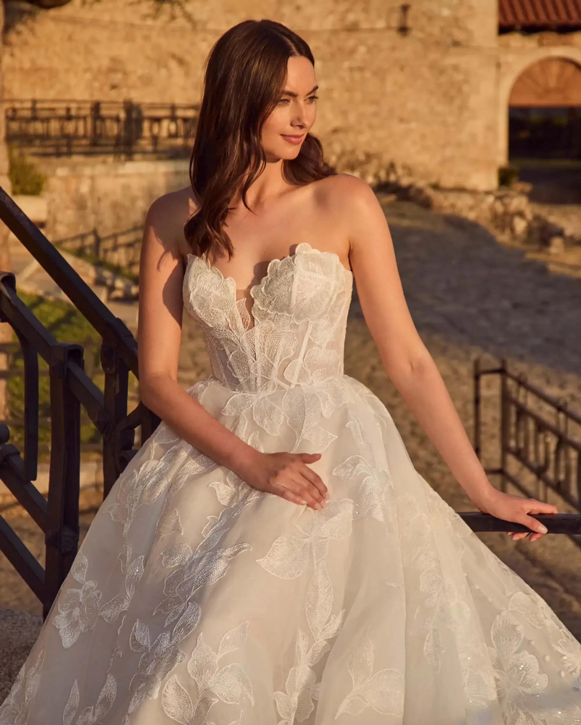 A woman in a strapless, floral lace wedding dress stands on stone steps, smiling softly. Sunlit medieval architecture creates a warm, romantic backdrop.