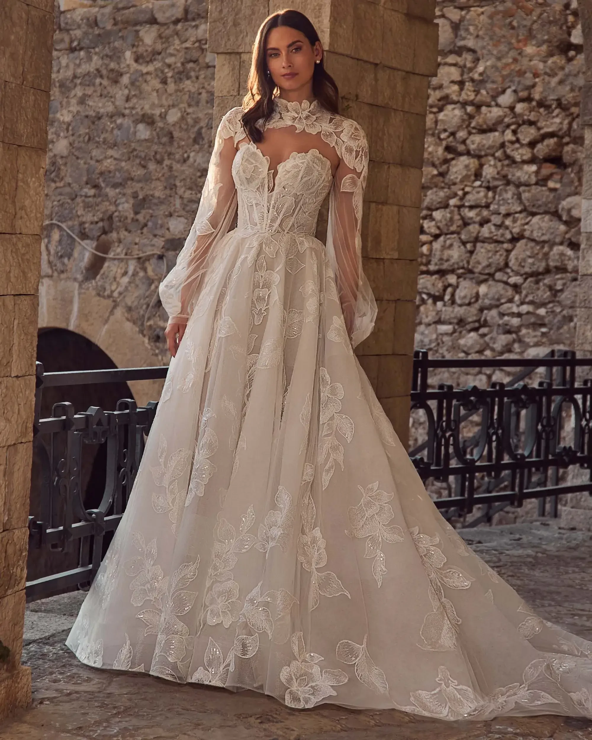 A woman in an elegant, floral lace wedding gown with sheer long sleeves stands by a stone wall. The scene evokes romance and timelessness.
