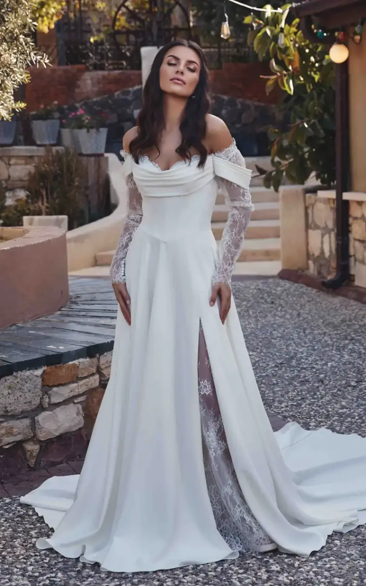 A woman in a white wedding dress with off-the-shoulder sleeves and a thigh-high slit, standing outdoors among greenery and stone paths.
