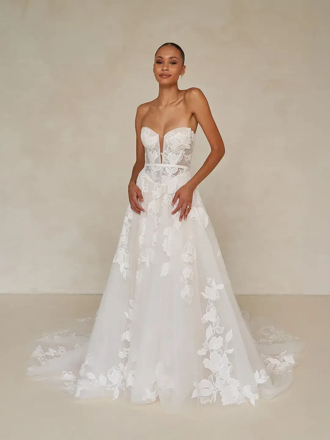 A bride stands confidently in a strapless, floral lace wedding gown with a flowing train. The backdrop is a simple, soft beige, enhancing elegance.