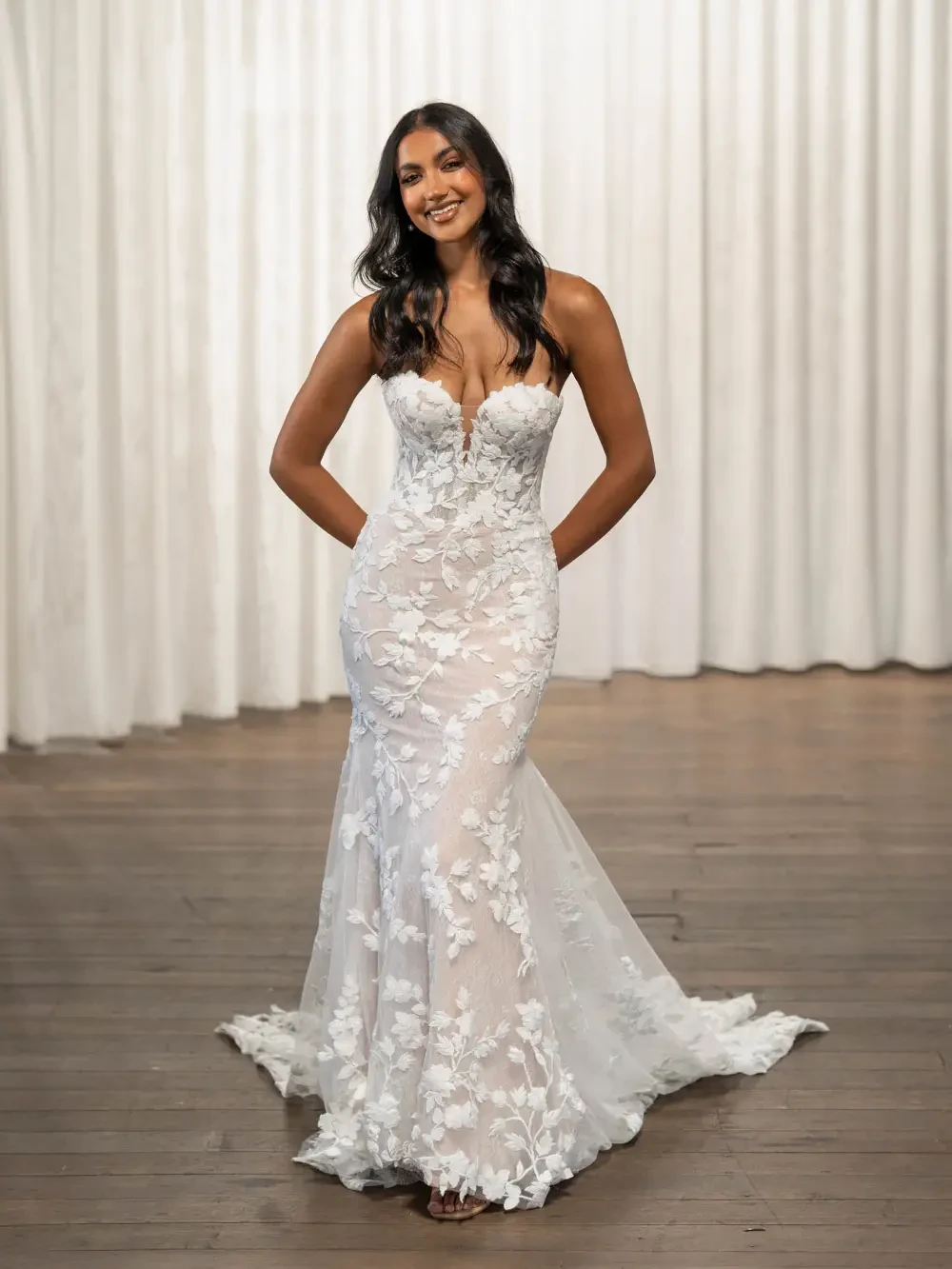 A woman in an elegant off-the-shoulder bridal gown adorned with floral lace patterns stands confidently. The soft focus background of white curtains adds a serene, romantic tone.