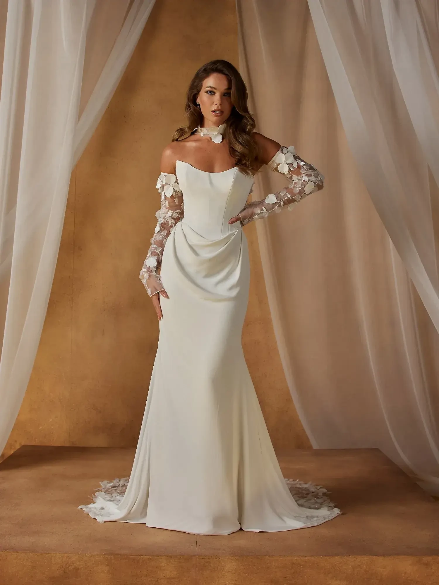 A woman wearing an elegant white off-the-shoulder dress with floral lace sleeves, standing gracefully against a warm-toned backdrop.