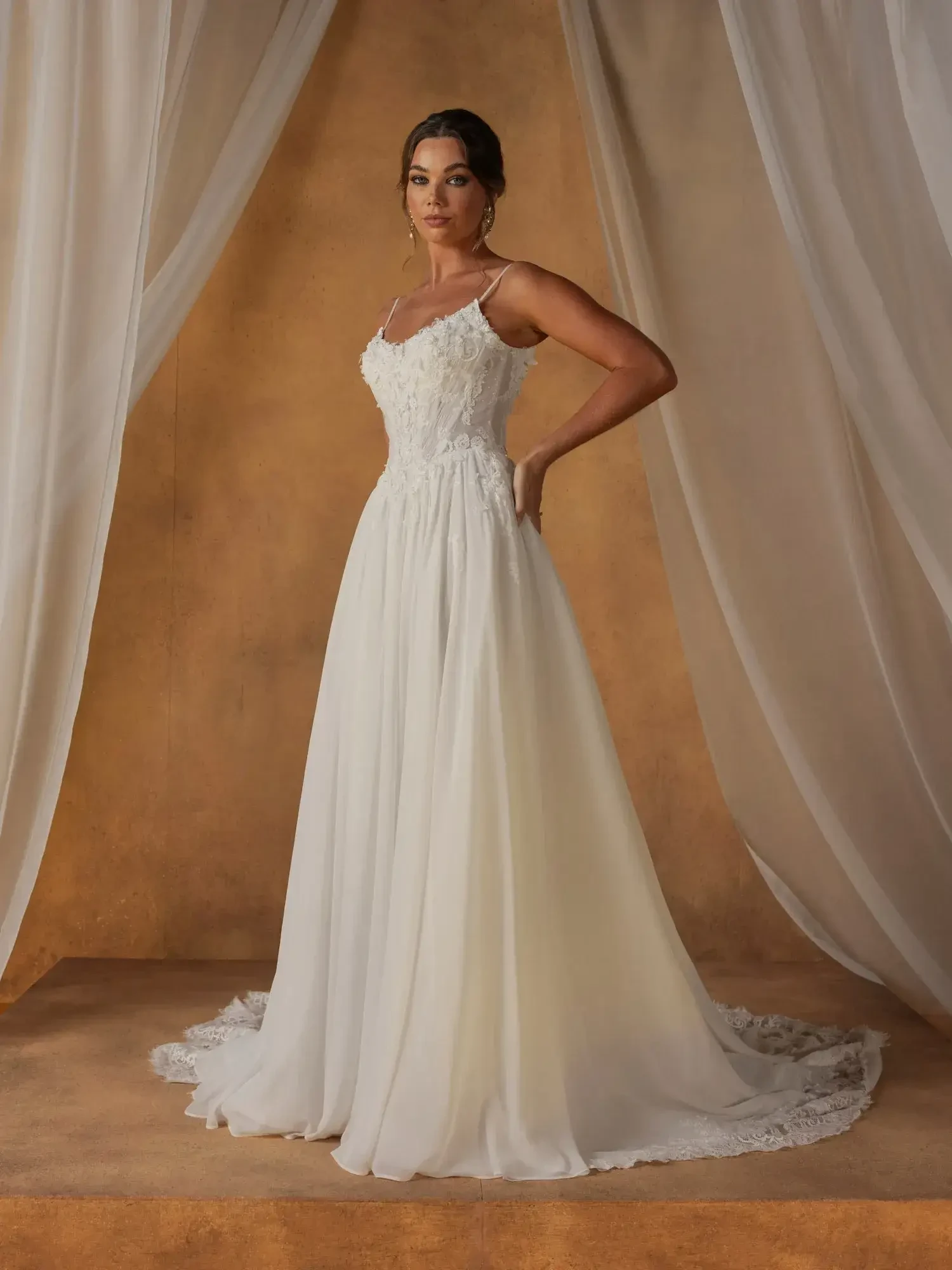 A model wearing a white wedding dress, standing elegantly against a soft backdrop with draped fabric.