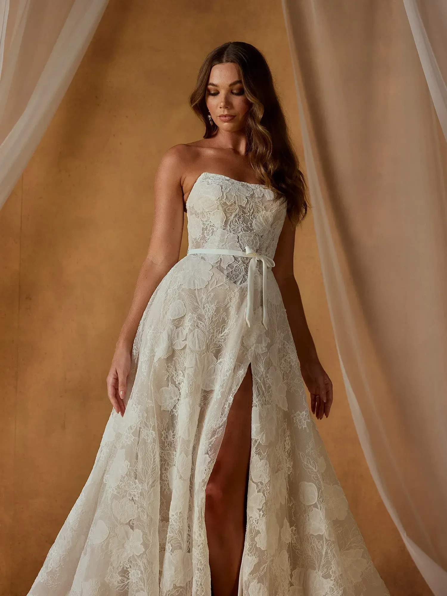 A woman in a strapless white lace dress with a high slit, posing in front of a light brown background draped with sheer fabric.