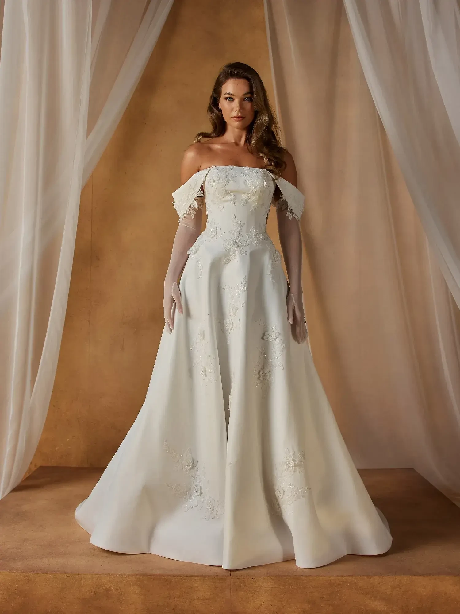 A woman stands elegantly in a white, off-the-shoulder, floral-embroidered gown. She is framed by sheer white curtains, exuding a graceful and serene vibe.