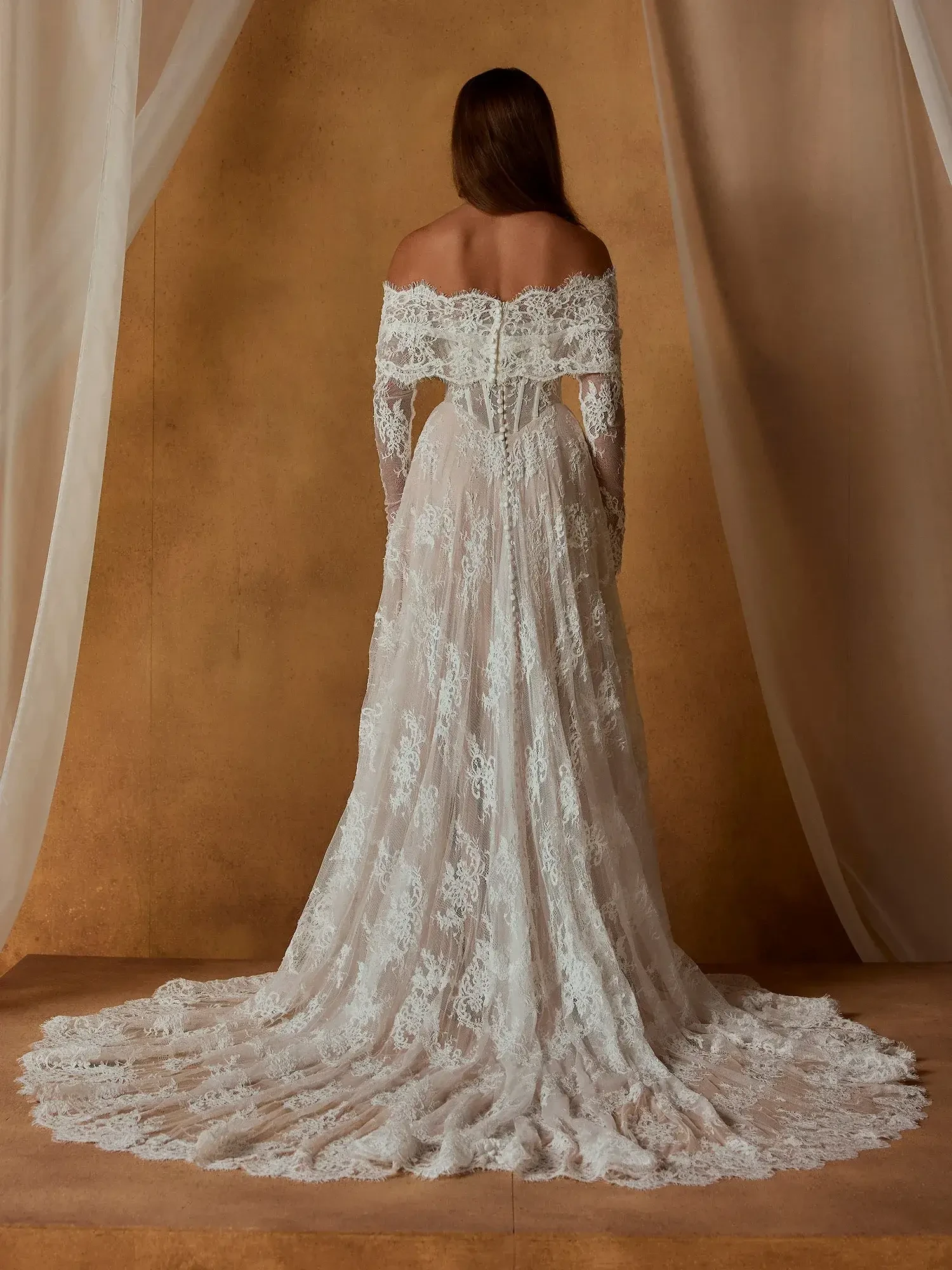 A woman wearing a long, off-the-shoulder lace wedding dress, standing with her back to the camera. The dress features intricate lace details and a train that flows on the floor. The background is a warm, neutral color with soft fabric draping.