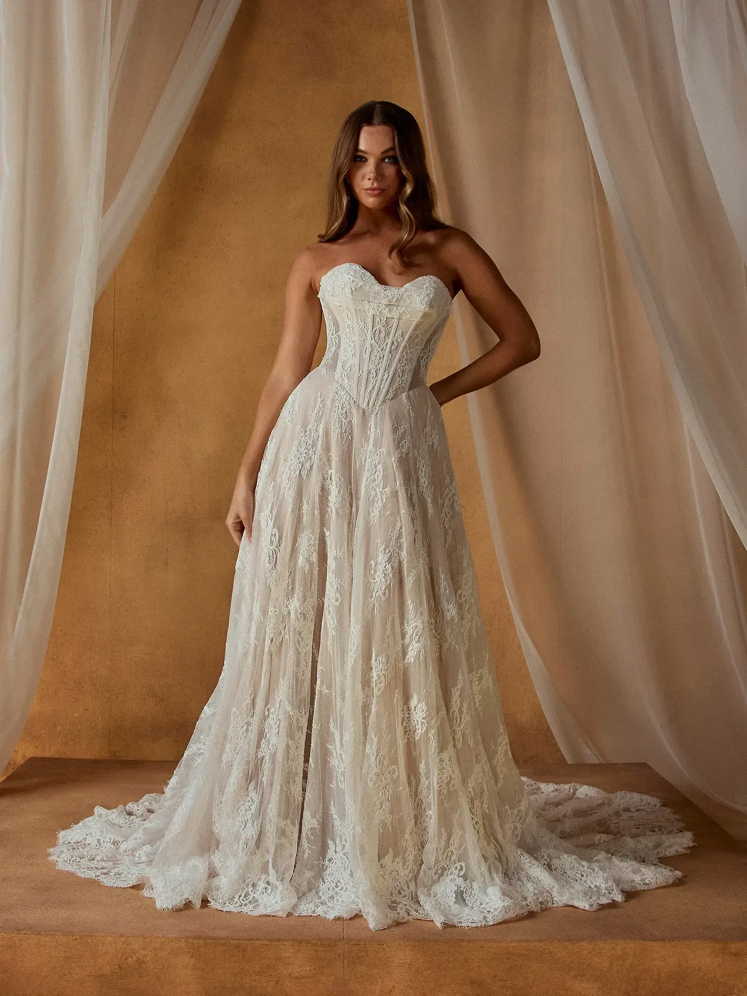 A woman in a strapless wedding dress with intricate lace detailing, standing against a soft, neutral background.