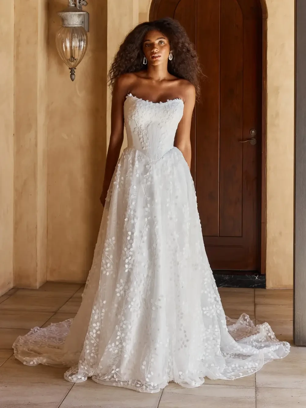 A model wearing a strapless white wedding dress with floral embellishments, standing indoors in front of a wooden door.