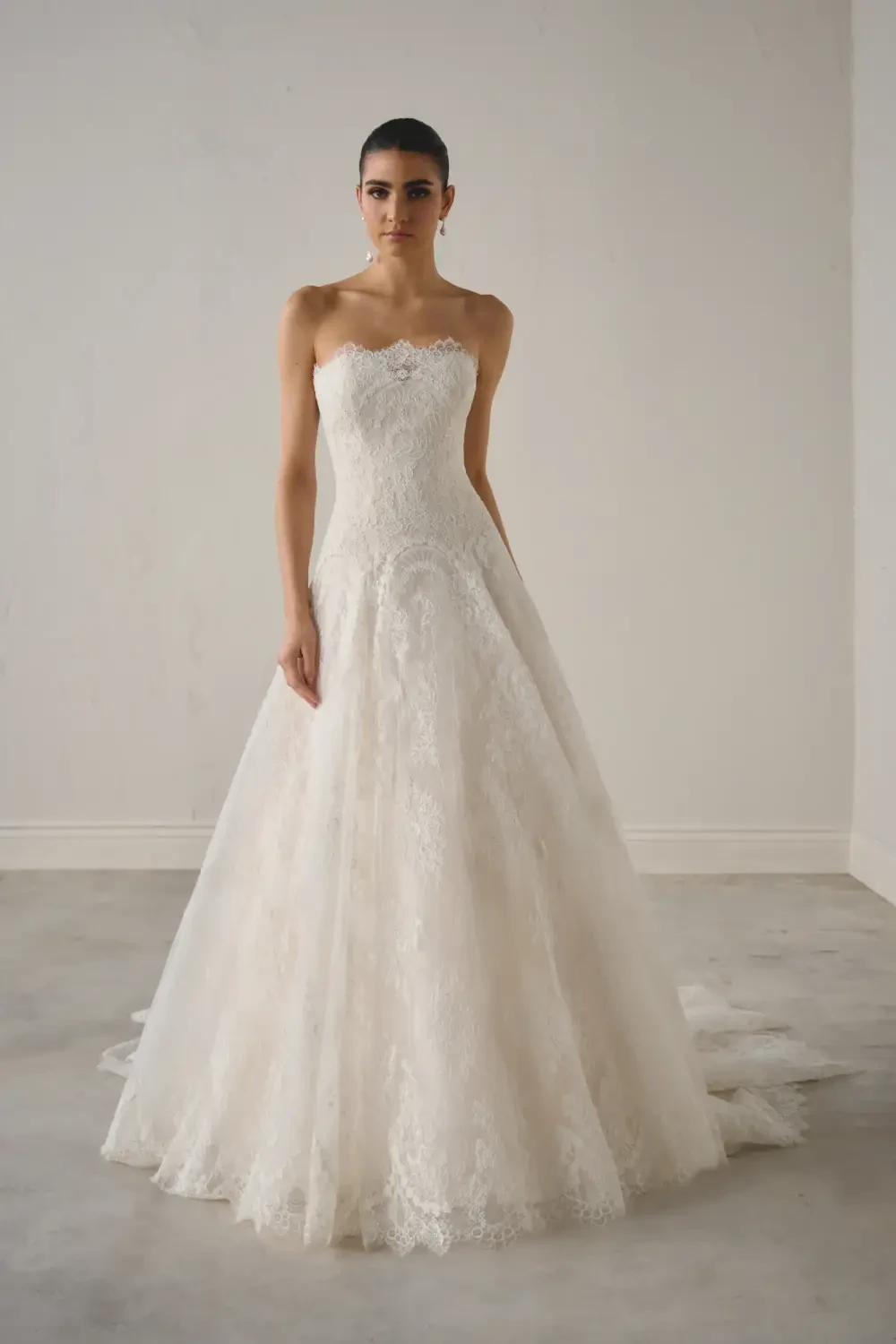 A woman wearing a strapless wedding dress with a lace bodice, standing in a bright, minimalistic room.